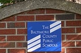 Blue and white sign on a red brick wall says, “The Baltimore County Public Schools.