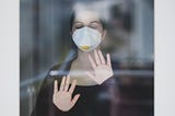 Photo of woman with face mask behind glass wall. Photo posted on Dr. James Goydos 2021 article about mask requirements and debate between Dr. Fauci and Rep. Jordan