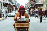 Photo of a person sitting in the middle of the street at a small wooden desk that says “Poet for hire” on the front. The person is using a typewriter that is on top of the desk.
