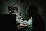 A person writing on a desk in dark room, only lit by a table lamp.