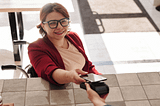 A smiling woman using a wheelchair holds her smartphone over a payment device at a cafe.