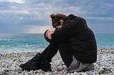 Person sitting on a rocky beach with their head down, representing the deep emotional struggle and self-blame that often follows trauma, as discussed in the article about overcoming the effects of trauma and shifting away from self-criticism.