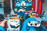 A room from a person Point of View with coffee mug that says — But first Coffee, then a gaming console, TV, and stuff toys of Snorlax in the background