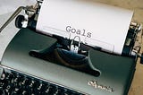 An antique typewriter on a wooden desk with a sheet of paper in it with “Goals” typed on it.