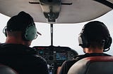 Pilot and co-pilot sitting in the cockpit with headphones on viewed from the back of their heads and out the cockpit window.