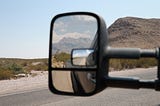 Car side view mirror with a mountain vista in view, with the road and more vistas visible behind the mirror in the direction the car is headed.