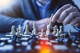 A faded upper chest of a man in a blue suit jacket and light blue shirt holding a white chess piece between two fingers over a full dark blue and white chess board of pieces. Writer Christine McDonald uses the picture to symbolize the strategy it takes to win.