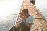 A father holds his daughter on a dock at the lake with the sun glistening behind them