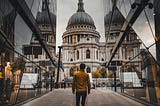 Image; man walking along a city street, wearing a short jacket and jeans, facing a church tower with his back to the viewer