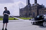 A man stands to the left of the picture. A disused old, large building is in the background. A black car is on the right of the picture. The man holds a long piece of paper that stems from the car.