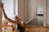A woman in a leotard bends over backwards toward her heels with her mouth open. She is in a sparsely-furnished hardwood floor house, bathed in natural light.