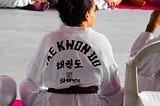 A student of TaeKwonDo, wearing a white uniform, sits on a mat with others looking towards their right.