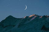 Mountain top early evening. Fingernail moon. Faint stars. The last of the sunlight is catching the peaks of the mountain.