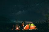 A picture of a group of campers at night enjoying a campfire. A green and orange tent that is lit from the inside sits beside them.