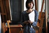 Young female artist looking pensive. She is standing in her studio holding her sketch pad. She looks like she is listening carefully to someone.