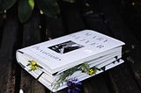 A snow white book sitting on a brown plank with tiny yellow and purple flowers sitting in between its pages.