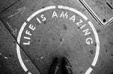 Image of a dark grey ground with a white circle painted on it with the worlds “Life Is Amazing” painted inside the circle. The image was chosen by Spiritual Lifestyle Writer The Christine McDonald to highlight how amazing life is when you work toward your physical goal alongside the guidance of your spiritual self.