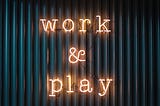 A black background featuring vertical black wood slats overlaid with a neon sign which has work & play illuminated.