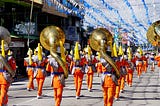 A marching band wearing orange outfits, carrying lots of tubas.