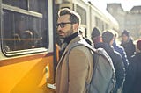 A man wearing glasses, a tan coat, and a grey backpack is standing near the door of a yellow bus. He looks slightly to his lrft, his expression intent, as people around him line up to board. The scene is bright with daylight, and thebus and crowd suggest a busy urban setting.