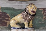 A ceramic lion on a marble mantle shelf. The lion is naively made with an almost human face, and glazed in with yellow body and face and brown mane. The glaze is chipped in places. Around its neck is a circle of small back beads. Behind the lion, the wall is roughly but richly painted in earthy green, brown, yellow, grey and white.