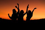 A dark shadow of three women rejoicing in front of a sunset.