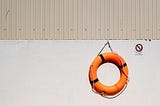 A bright orange lifebuoy hanging from a rope against a cream colored wall.