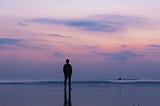 A man standing on the beach looking out at the waves at sunset.