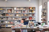 A brightly lit bookstore, with white shelves and a white table, both full of books for sale.