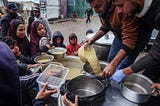 Two men pour lentils into a pot from a large vat while children circle them with their containers outreached, waiting for their share.