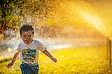 A little boy running with a smile on his face while a sprinkler is spraying water in his direction. The drops of water are golden because the sun is hitting them