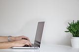 Two hands typing on a laptop, which sits on a white table. The picture is taken with the hands and laptop sideways, to the lefthand side of the picture. A green plant is slightly off frame on the right hand side