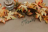 A wreath of dried leaves with a candle in the middle and the words “give thanks” written on the table
