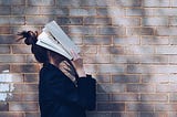 Uma mulher com um livro cobrindo o rosto sobre uma parede de tijolos.