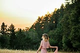 A pretty blonde girl in a long pink flown dress dances in a golden grass field on the edge of a wooded forest at sunset.