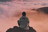 A man sitting on a mountain peak and staring at the clouds at sunset