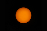 Image of the sun taken through an amateur telescope. The surface is perfectly round and orange. A few sunspots are visible as faint black smudges.