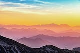 an alluring landscape of dusky mountains with a person standing, pinkish-yellow horizon and white/blue skies.