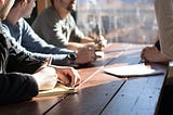 An image of 4 people sitting around a table. You can see their hands, some are writing on paper. You don’t see their faces clearly.