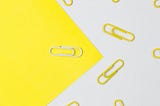 Piece of yellow paper on an angle with one white paperclip on it against a white background with many yellow paperclips on it