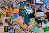 a woman lies on a bed with books all around her and on top of her. one open at eye level she’s engrossed in.