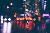 Imagem noturna do trânsito de uma cidade, mostrando ao fundo luzes acesas coloridas e desfocadas