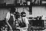 Two men, workers in a restaurant, with aprons on are in a kitchen preparing food.