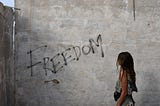 Lightly black spray-painted word “FREEDOM” on a concrete wall being stared at contemplatively by a young women whose face is hidden by her long hair giving the sense more of her staring at the meaning of this word.