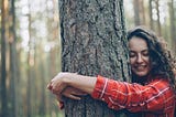 a woman in a red plaid shirt encircles a tree trunk iwth her arms and presses her face against the bark
