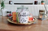 A picture of a glass jar covered with a white paper with the words “gratitude jar” amidst other deco
