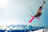 airborne skiier crosses red skis midair with snowy countains in background