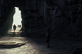 A distant figure strides out of an ancient rock cave towards a crack that leads out towards the sun.