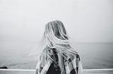 A black and white image of a woman facing the ocean with wind in her hair