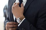 Image of a man in a suit, where you can only see part of his torso, one hand straightening his tie, and on that wrist is a watch with a brown wristband.